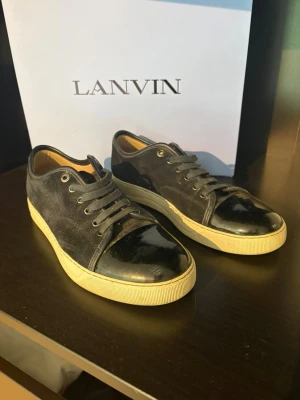 Svarta Lanvin sneakers i mocka - Snygga svarta sneakers från Lanvin med ovandel i mocka och lackad tå. Skorna har vita sulor och klassisk låg profil med svarta skosnören. Däremot ser sulan gul ut men det är endasr ljuset. Skicka iväg ett medelande om du vill ha fler bilder eller diskutera pris!