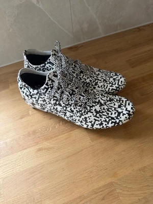 Under armour  fotbollsskor med mönster - Unika under armour  fotbollsskor med svartvitt pixelmönster och snörning. Skorna har låg profil och dobbar för bra grepp på planen. Tillverkade i syntetmaterial för lätt känsla och smidig passform. Perfekta för dig som vill sticka ut på fotbollsplanen.