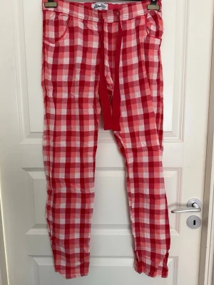 Rödrutiga pyjamasbyxor från MaxMart - Säljer ett par pyjamasbyxor från MaxMart i rött och vitt rutigt mönster. Byxorna har resår i midjan, dragsko och två sidofickor. Tillverkade i ett mjukt och lätt tyg, perfekta för att chilla hemma. Liten fläck på höger byxben, se bild. 