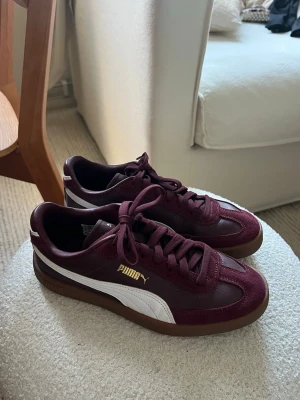 Vinröda Puma sneakers med vit detalj - Snygga vinröda sneakers från Puma med klassisk vit sidostrip och guldfärgad logga. Skorna har en lägre brun gummisula och kombinerar mocka och läder för en stilren look! Införskaffade mig dessa 2025 men dom är knappt använda och därför väldigt bra skick! Ordinarie pris: 799:-