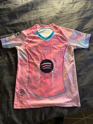 Barcelona rosa matchtröja Nike - Snygg Barcelona matchtröja i rosa och ljusblå toner med klubbmärke, Nike-logga och Spotify som sponsor på bröstet. Tröjan har korta ärmar, V-ringning med blå kant och coola grafiska detaljer. Perfekt för dig som älskar fotboll och vill sticka ut på planen. Säljer då den inte passade