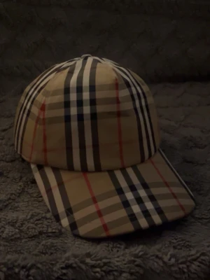 Beige keps från Burberry med rutmönster - Säljer en klassisk beige keps från Burberry med det ikoniska rutmönstret i svart, vitt och rött. Kepsen har böjd skärm och justerbar rem bak. Märkt med 'BURBERRY LONDON ENGLAND' på baksidan. Perfekt för dig som gillar stilrena accessoarer.