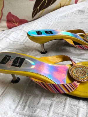Gula sandaler med holografisk sula - Unika gula sandaler från Sarah Zanka med holografisk sula, smala remmar i rosa och vitt samt rund dekoration med små glittriga stenar. Klack i metall och öppen tå ger en cool och färgstark look. Perfekta för dig som vill sticka ut. Väldigt lite använda men mindre skavanker som syns på bilderna