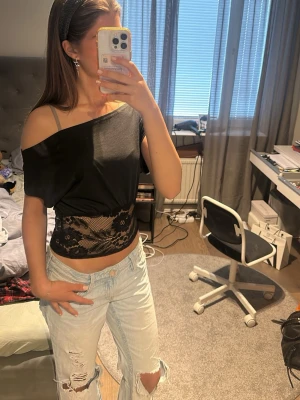 Svart croppad topp med spetsdetalj - Trendig svart croppad topp med offshoulder-design och bred spetsdetalj i midjan. Toppen är ärmlös på ena sidan och har en lös passform som ger en avslappnad vibe. Perfekt för dig som vill sticka ut med en unik och snygg topp.