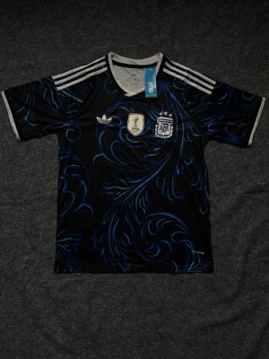 Argentina Adidas fotbollströja - Snygg svart fotbollströja från Adidas med blått mönster och vita detaljer. Tröjan har Argentinas landslagsemblem, FIFA-märke och tre vita ränder på axlarna. Perfekt för dig som älskar fotboll och vill sticka ut på planen. Finns i S, M och L