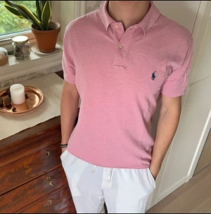 Rosa pikétröja från Ralph Lauren - Säljer en klassisk rosa pikétröja från Ralph Lauren med korta ärmar och den ikoniska loggan broderad på bröstet. Tröjan har krage och knappar vid halsen, perfekt för en clean och stilren look.