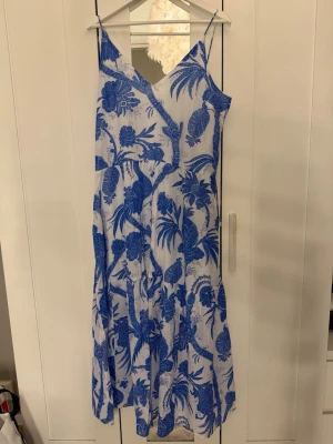 Blåvit klänning med blommigt mönster - Säljer en somrig blåvit klänning med tunna axelband och v-ringning. Klänningen har ett iögonfallande blommigt mönster i blått på vit botten och är i en luftig, lätt form. Perfekt för varma dagar och semester.