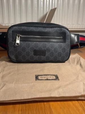 GG svart shoppingväska med monogram - Snygg svart shoppingväska från GG med klassiskt GG-monogram i grått. Väskan har en framficka med dragkedja och justerbar axelrem i blått och rött. Tillverkad i slitstarkt canvasmaterial med läderdetaljer. Perfekt för dig som vill ha en trendig och praktisk väska.