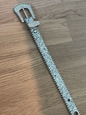 Glittrigt silverbälte med nitar - Säljer ett riktigt snyggt bälte i silver med glittriga stenar och nitar längs kanterna. Spännet är dekorerat med stenar för extra bling och bältet har flera metallhål. Perfekt för dig som vill sticka ut och addera lite extra till din outfit.