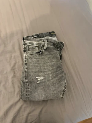 Grå slitna skinny jeans från Jack & Jones - Säljer ett par grå skinny jeans från Jack & Jones med slitningar och distressed detaljer på benen. Jeansen har klassisk femficksmodell, bälteshällor och dragkedjegylf. Perfekta för en avslappnad och trendig look.