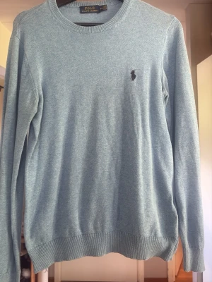 Ljusblå stickad tröja Polo Ralph Lauren - Ljusblå stickad tröja från Polo Ralph Lauren med rund halsringning och diskret broderad logga på bröstet.  Storlek m