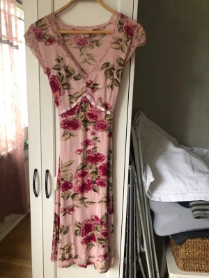 En blommönstrad Midsommarklänning i rosa toner - Romantisk långklänning i ljusrosa med stora blommor i rosa och grönt. Perfekt till midsommarfesten! Klänningen har v-ringning, spetsdetaljer vid axlarna och ett knytband i midjan för en feminin siluett. Finns ett (eller ett par) pyttelitet hål, se bild. Detta drar förstås ner priset en del. I övrigt en fantastisk klänning, använd endast vid ett tillfälle.