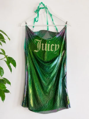 Grön glittrig klänning Juicy Couture - Grön glittrig kortklänning från Juicy Couture med halterneck och texten 'Juicy' framtill. Klänningen har en skimrande metallic-finish och tunna band som knyts i nacken. Perfekt för dig som vill sticka ut och glänsa.