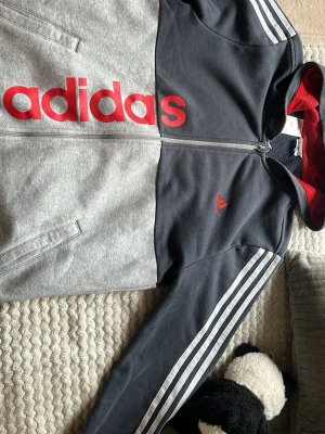 Grå och svart hoodie från Adidas - Snygg hoodie från Adidas i grått och svart med röd logga och detaljer. Dragkedja framtill, två fickor och klassiska vita ränder på ärmarna. Insidan av huvan är röd med meshfoder. Perfekt för en sportig och avslappnad stil.