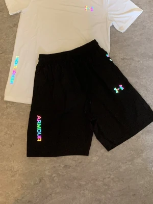 Under Armour träningsset, vit/svart - Snyggt träningsset från Under Armour med en vit t-shirt och svarta shorts. Setet har regnbågsfärgade loggor och textdetaljer. Perfekt för gymmet eller löpning. Materialet är lätt och andas, vilket gör det bekvämt under träning.
