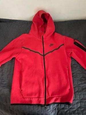 Röd Nike hoodie med dragkedja - Snygg röd hoodie från Nike med hel dragkedja framtill och svart Nike-logga på bröstet. Hoodien har svarta detaljer längs bröstet och en ficka med dragkedja på ena ärmen. Tillverkad i mjukt bomullsmaterial och har två fickor fram samt huva.