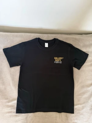 Svart Gallery Dept t-shirt - Snygg svart t-shirt från Gallery Dept. med gul logga och text på bröstet. Klassisk rund hals och korta ärmar. Tillverkad i mjuk bomull som känns skön mot huden. Perfekt för dig som gillar streetwear och vill ha en enkel men stilren look.