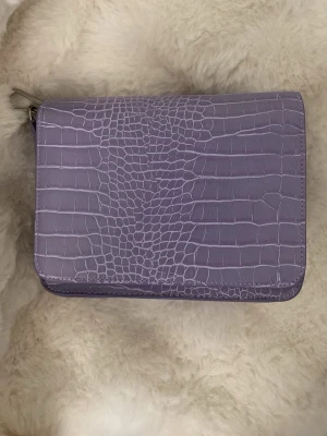 Ljuslila axelväska med krokoprägling - Säljer en ljuslila axelväska i fyrkantig modell med snygg krokodilpräglad yta. Väskan har ett stilrent och modernt utseende, perfekt för dig som gillar trendiga accessoarer. Materialet är konstläder och väskan har metallfäste för axelrem.
