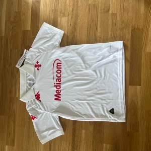 Vit Fiorentina fotbollströja Kappa S - Snygg vit Fiorentina fotbollströja från Kappa i storlek S. Tröjan har röd Mediacom-logga på bröstet, röda Kappa-loggor på axlarna och texten 'Fiorentina' i nacken. Tillverkad i lätt och ventilerande polyester, perfekt för match eller träning.