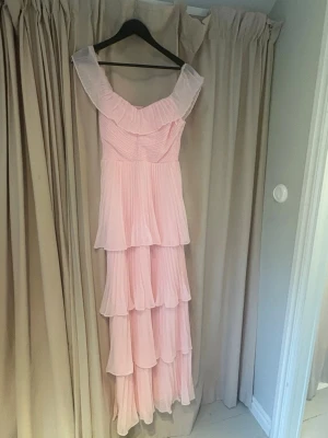 Balklänning, Ljusrosa långklänning med volanger - Elegant ljusrosa långklänning(helt ny, oanvänd med prislapp kvar)med flera lager plisserade volanger och offshoulder-design. Klänningen har en romantisk och flowy siluett som ger en drömmig känsla. Perfekt för speciella tillfällen där du vill sticka ut.