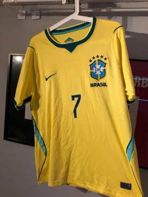 Brasilien matchtröja Vini Jr. #7 Nike - Säljer en gul Brasilien matchtröja från Nike med gröna detaljer och nummer 7. På bröstet finns CBF-emblemet och Nike-loggan, och på ryggen står det VINI JR. samt nummer 7. Tröjan har korta ärmar och är tillverkad i ett lätt, ventilerande material.