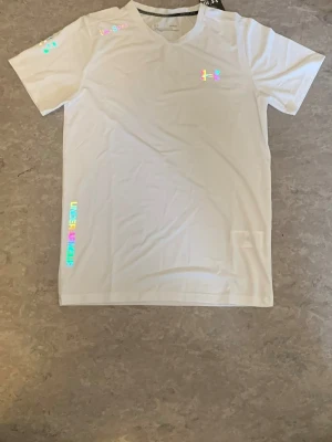 Under Armour träningsset, vit/ - Säljer ett snyggt träningsset från Under Armour med en vit t-shirt och . Båda plaggen har holografiska loggor och textdetaljer. T-shirten har rund hals och kort ärm, shortsen har resår i midjan. Perfekt för gymmet eller löpning.
