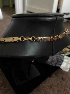 Guldigt halsband med länkdesign - Snyggt halsband i guldfärgad metall med unik länkdesign. Halsbandet har rektangulära och dekorativa länkar som ger ett stilrent och trendigt intryck. Perfekt för dig som vill ha ett statement-smycke som sticker ut och kompletterar din outfit.