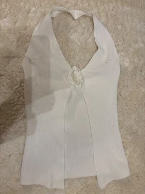 Vit ribbad halterneck topp med rosdetalj - Säljer en vit ribbad halterneck topp med en snygg rosettliknande rosdetalj framtill. Toppen är ärmlös och har en djup v-ringning som knyts i nacken. Materialet är stretchigt🌸