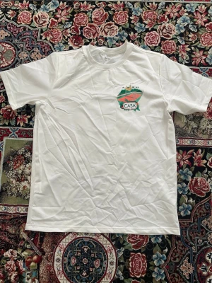 Vit CASA BLANCA t-shirt med tryck - Säljer en vit t-shirt från CASA med färgglatt tryck både fram och bak. Trycket visar en illustration med gröna, röda och gula detaljer samt texten CASA. T-shirten har rund hals och korta ärmar, perfekt för en avslappnad stil.