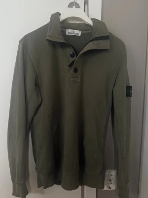Olivgrön sweatshirt från Stone Island - Snygg olivgrön sweatshirt från Stone Island med klassisk patch på ärmen. Tröjan har hög krage med 4 knappar och dragkedja. Strl M