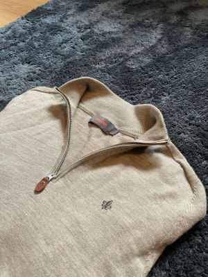 Beige halvzip-tröja från Morris - Snygg beige stickad tröja från Morris med halvzip och diskret broderad logga på bröstet. Tröjan har långa ärmar, ribbade muddar och är tillverkad i mjukt material som känns skönt mot huden. Perfekt för lager-på-lager och stilren look. Tröjan är liten i storlek och passar som S(se mått). Det finns ett litet hål som inte syns när man bär tröjan.(På ryggen).