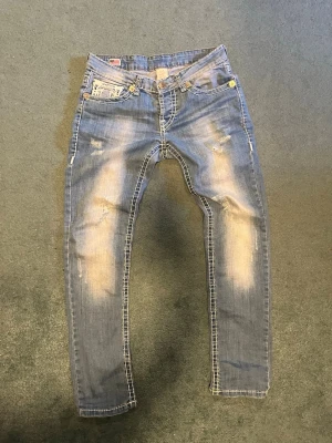 Ljusblå distressed jeans från True Religion - Säljer ett par ljusblå jeans från True Religion med slitna detaljer och kontrastsömmar. Jeansen har broderad logga på fickan och klassisk femficksmodell. Perfekta för en avslappnad och trendig look.