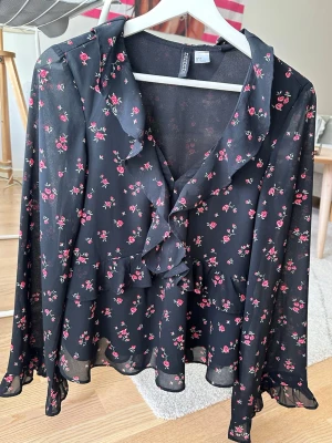 Svart blommig volangblus - Söt svart blus från H&M med små röda rosor och volangdetaljer längs urringningen och framtill. Har inte använts på flera år och är i väldigt bra skick. Perfekt för en trendig och romantisk look.