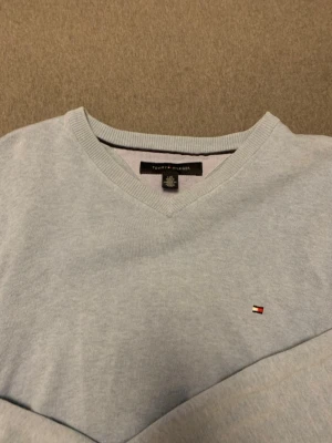 Ljusblå stickad tröja Tommy Hilfiger - Ljusblå stickad tröja från Tommy Hilfiger med V-ringning och diskret logga broderad på bröstet. Tröjan har långa ärmar och ribbade muddar vid ärmslut och nederkant. Perfekt för lager på lager och enkel att matcha.