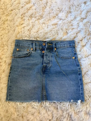 Blå jeanskjol med fransig kant - Säljer en blå jeanskjol från Lager 157 med fransig rå kant nertill och klassiska fem fickor. Kjolen har normal passform, bälteshällor och stängs med knapp och dragkedja framtill. Perfekt för dig som gillar en avslappnad och trendig stil.
