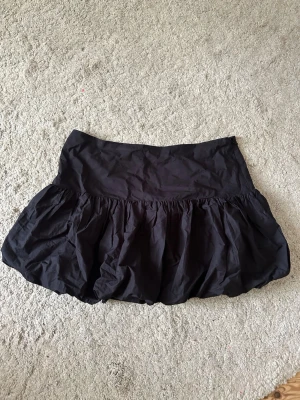 Svart ballongkjol - Säljer en svart minikjol med volang och ballongformad siluett. Kjolen har en bred midja och är gjord i ett lätt, prassligt material som ger extra volym. Perfekt för dig som vill ha en statement piece i garderoben.
