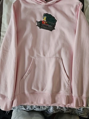Ljusrosa hoodie med Simpsons-brodyr - Säljer en ljusrosa hoodie med en broderad bild av Mr. Burns från Simpsons utklädd till Dracula på bröstet. Hoodien har huva och magficka, samt ribbade muddar vid ärmslut och nederkant. Perfekt för dig som gillar unika och roliga tryck!