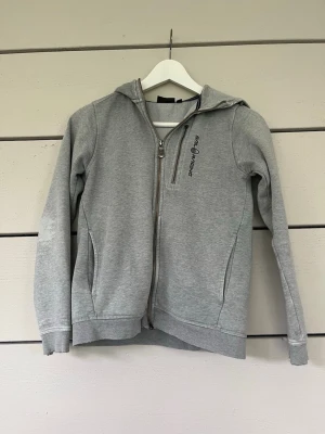 Grå zip-hoodie från Sail Racing - Snygg grå hoodie från Sail Racing med dragkedja framtill och två fickor. Hoodien har en broderad logga på bröstet och en liten detalj på huvan. Tröjan är lite sliten vid arm-mudden och armbågarna.