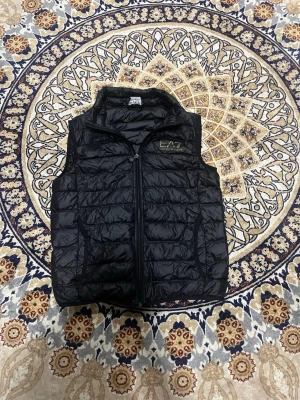 Armani väst S - Snygg svart dunväst från EA7 Emporio Armani med quiltad design och hög krage. Västen har dragkedja framtill och två sidofickor. Liten EA7-logga i guld på bröstet ger en exklusiv touch. Perfekt att slänga över en hoodie eller tröja för extra värme.