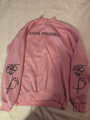 Rosa BTS Love Yourself sweatshirt - Rosa sweatshirt med texten 'LOVE YOURSELF' framtill och stor BTS-logga på ryggen. Svarta ros-motiv på båda ärmarna. Tröjan har ribbade muddar och rund hals. Perfekt för dig som älskar K-pop och vill ha en snygg och bekväm tröja.