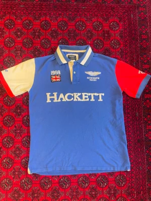 Blå Hackett Aston Martin Racing piké - Snygg blå pikétröja från Hackett x Aston Martin Racing med vita och röda kontrastärmar. Stor logga framtill och broderade detaljer som brittisk flagga och racingtext. Klassisk krage med knappar och sportig look.