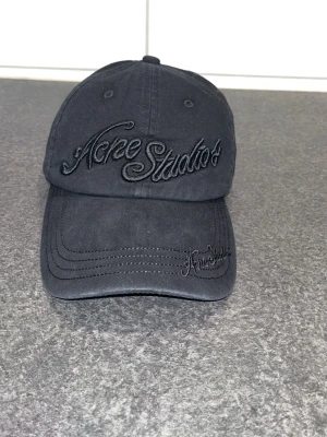 Svart keps från Acne Studios - Säljer en svart keps från Acne Studios med broderad logga framtill och på skärmen. Klassisk böjd skärm och justerbar baksida. Tillverkad i bomullstyg med snygga sömdetaljer. Perfekt accessoar för en clean och stilren look.
