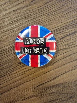 Punks Not Dead pinsknapp - Cool knapp med texten 'Punks Not Dead' i svart graffiti-stil. 4,4 cm. Perfekt accessoar för dig som gillar punk och vill sticka ut. Rund form och klassisk punkkänsla.