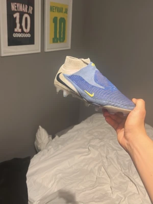 Nike phantom elite fotbollskor - Säljer ett par blåa Nike fotbollsskor med strumpliknande skaft och utan snörning. Skorna har gula detaljer och en svart Nike-logga på sidan. Ovandelen är i ett stickat material för bra passform och grepp på planen. Perfekta för dig som vill ha stilrena och moderna skor.