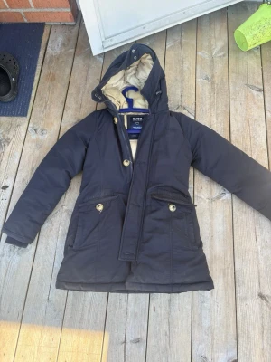 Svart dunjacka med huva från Svea - Säljer en svart dunjacka från Svea med beige foder och huva. Jackan har långa ärmar, guldiga knappar och två stora fickor framtill. Perfekt för kalla dagar och har en klassisk, normal passform. Hängare i nacken och snygga detaljer som gör den extra stilren.