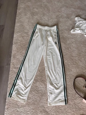 Adidas byxor  - Säljer ett par vita mjukisbyxor från Adidas med klassiska gröna ränder längs sidorna och Adidas-logga på låret. Byxorna har resår i midjan och raka ben. Perfekta för en avslappnad och sportig stil. Byxorna är knappt använda 