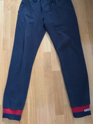 Mörkblå mjukisbyxor från Moncler - Snygga mörkblå mjukisbyxor från Moncler med elastisk midja och dragsko. Byxorna har fickor fram och röda detaljer vid bensluten där Moncler-loggan syns. Materialet är mjukt och bekvämt, perfekt för chill dagar. Klassisk Moncler-patch baktill.