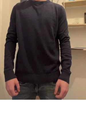 Mörkblå stickad tröja - En enkel och stilren mörkblå stickad tröja med rund halsringning och långa ärmar. Tröjan har ribbade muddar vid ärmslut och nederkant, vilket ger en snygg passform. Perfekt att matcha med jeans för en avslappnad look.