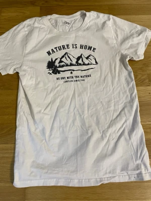 Vit t-shirt med naturtryck - Vit t-shirt med svart tryck av berg, träd och texten 'Nature is Home'. Skön passform och klassisk rund halsringning. Perfekt för dig som gillar outdoor-vibbar och enkel stil.