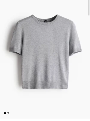 Grå kortärmad stickad topp - En stilren grå stickad topp med rund hals och korta ärmar. Toppen har ribbade kanter vid ärmslut och nederkant för en clean look. Perfekt att matcha med både jeans och kjol.